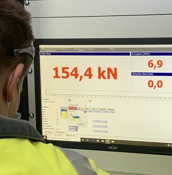 Eine Auszubildende sthet vor einem Monitor. Sie prüft den Druck, der auf einen Betonwürfel im Labor ausgeübt wird. Das Display zeigt 154,4 kN.