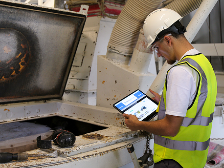Ein Auszubildender in Schutzausrüstung (PSA) kontrolliert mit einem Tablet in der Hand etwas an einem stationären Betonmischer