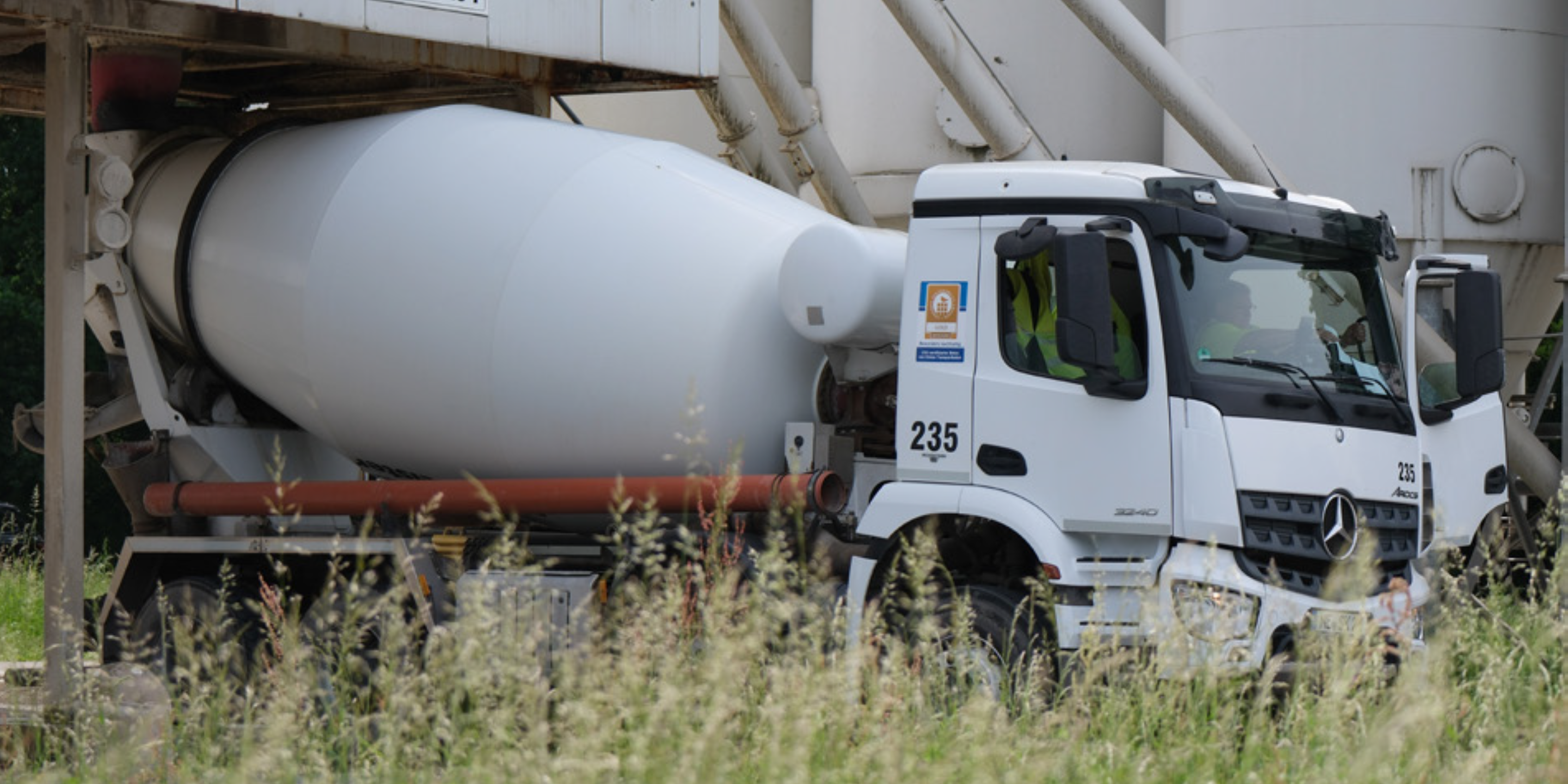 Betonmischfahrzeug beim Beladen an einer Betonmischanlage, teilweise verdeckt von hohem Gras im Vordergrund 