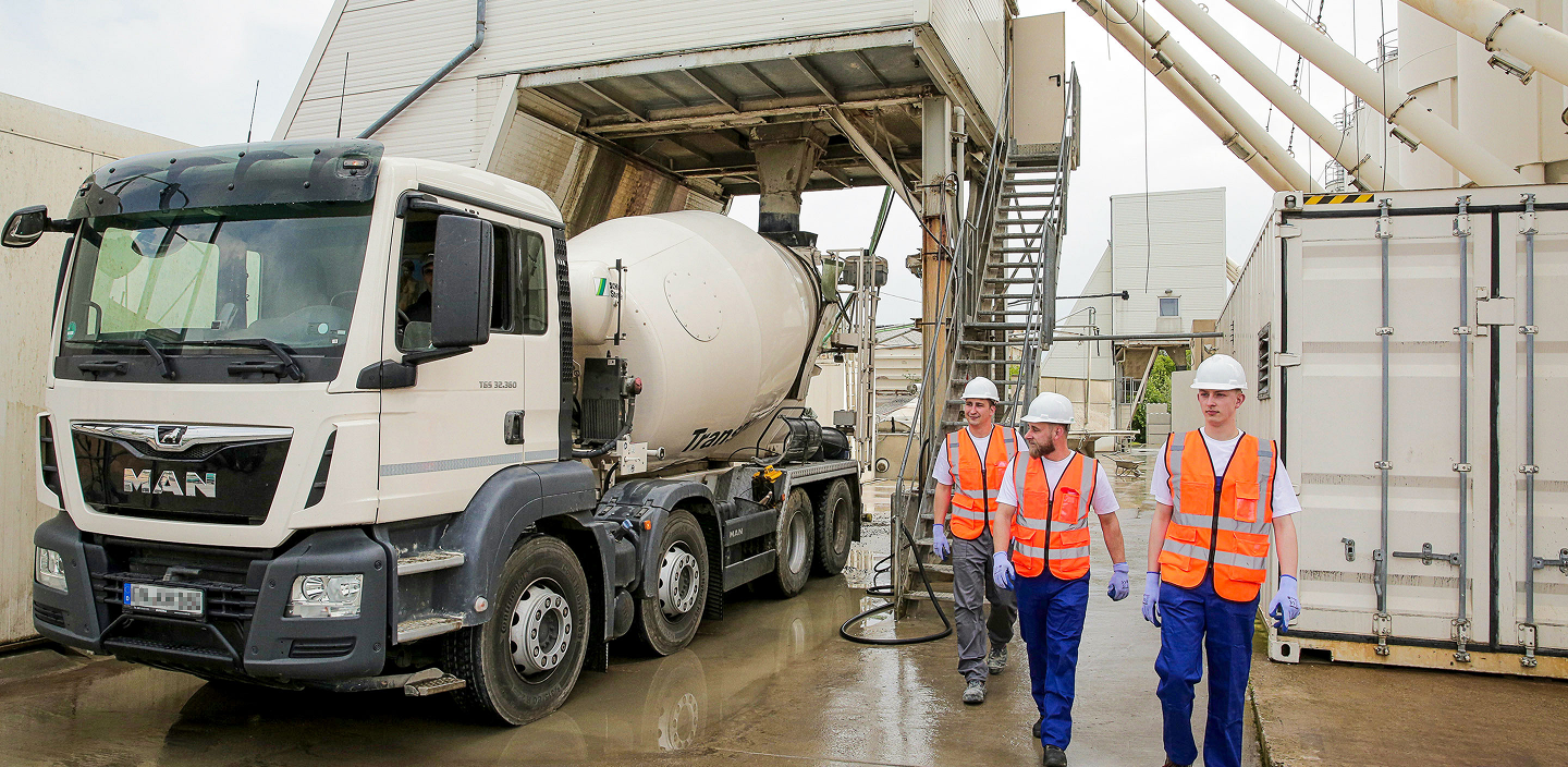 Drei Verfahrensmechaniker mit Helm und Warnweste gehen auf dem Gelände eines Betonwerks an einem Fahrmischer vorbei, der unter einer Befüllungsanlage steht.