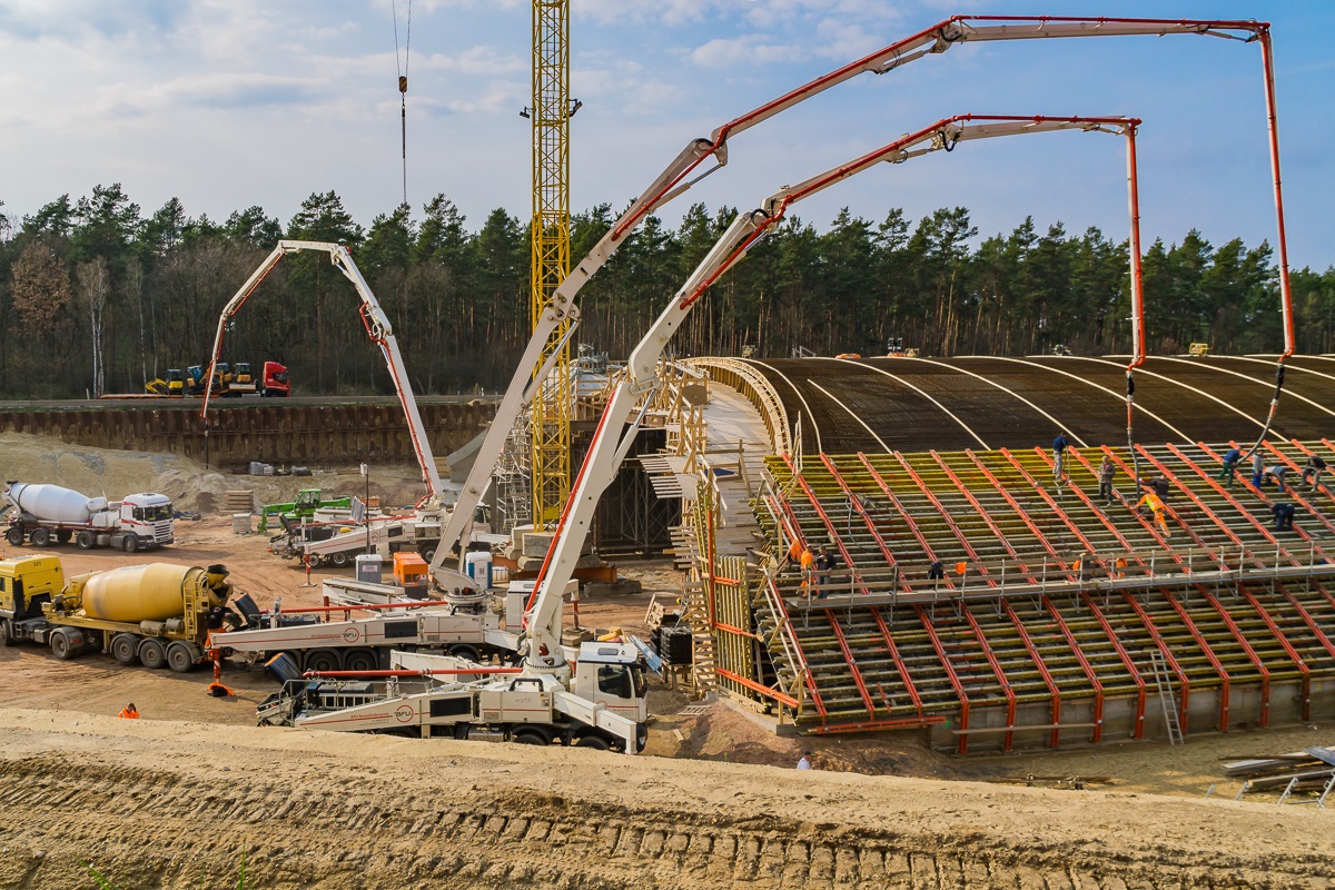 Mehrere Betonpumpen und Baufahrzeuge beim Betonieren einer großen Brücken- oder Tunnelkonstruktion auf einer Baustelle im Waldgebiet.
