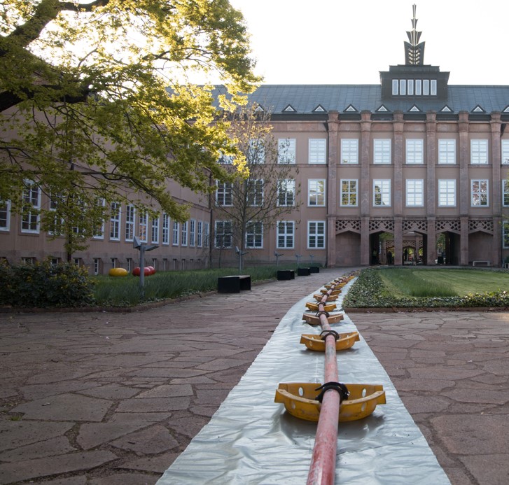 Langer Förderschlauch einer Betonpumpe verläuft über einen Innenhof auf ein mehrstöckiges repräsentatives Verwaltungsgebäude zu, im Vordergrund gepflasterter Platz.
